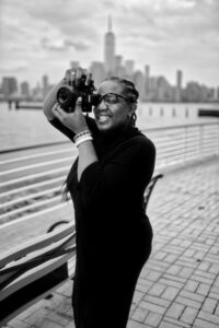 A black and white photo of a black woman photographer holding a camera to her eye with the NYC skyline in the background