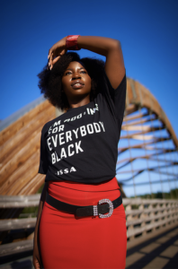 A blackwoman standing in a bridge in sunlight wearing a black t-shirt that says "I'm rooting for everybody black. -Issa""