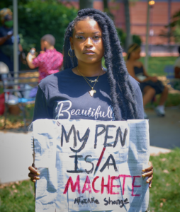 A black woman holding a handwritten sign that says My Pen is a Machete