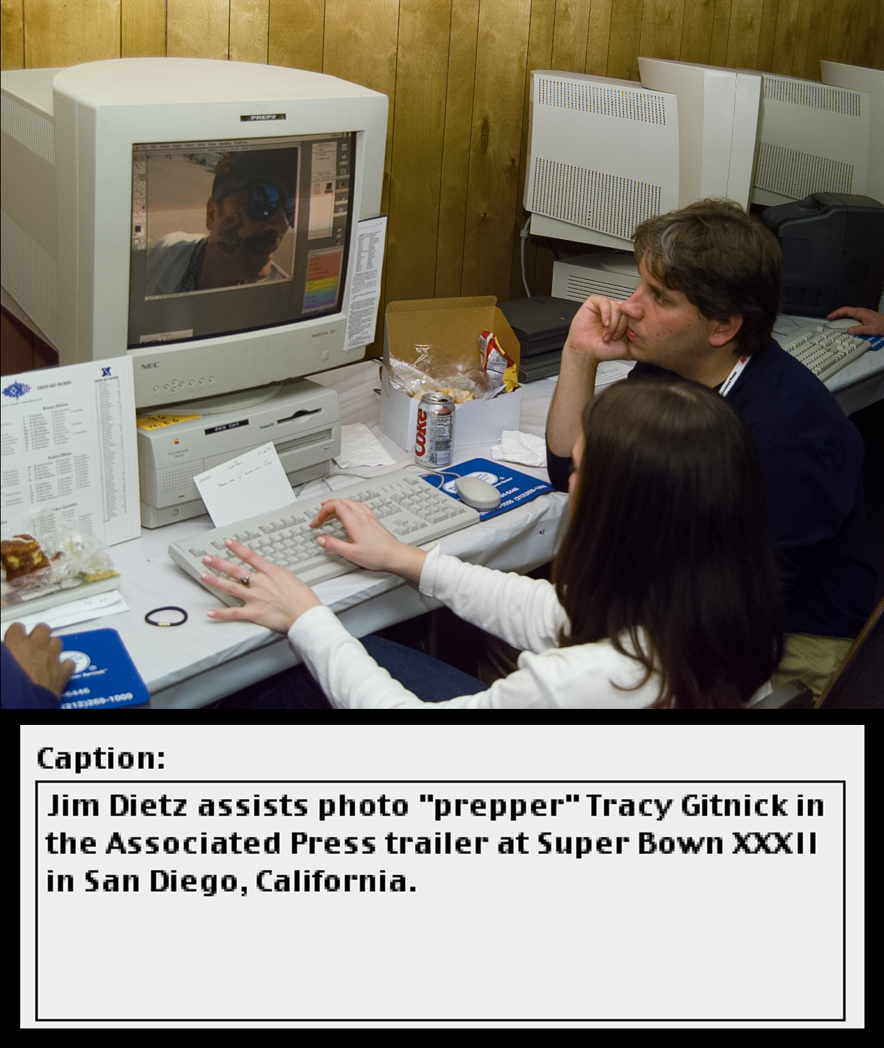 Two people sit at a computer workstation reviewing a photograph of a man in sunglasses