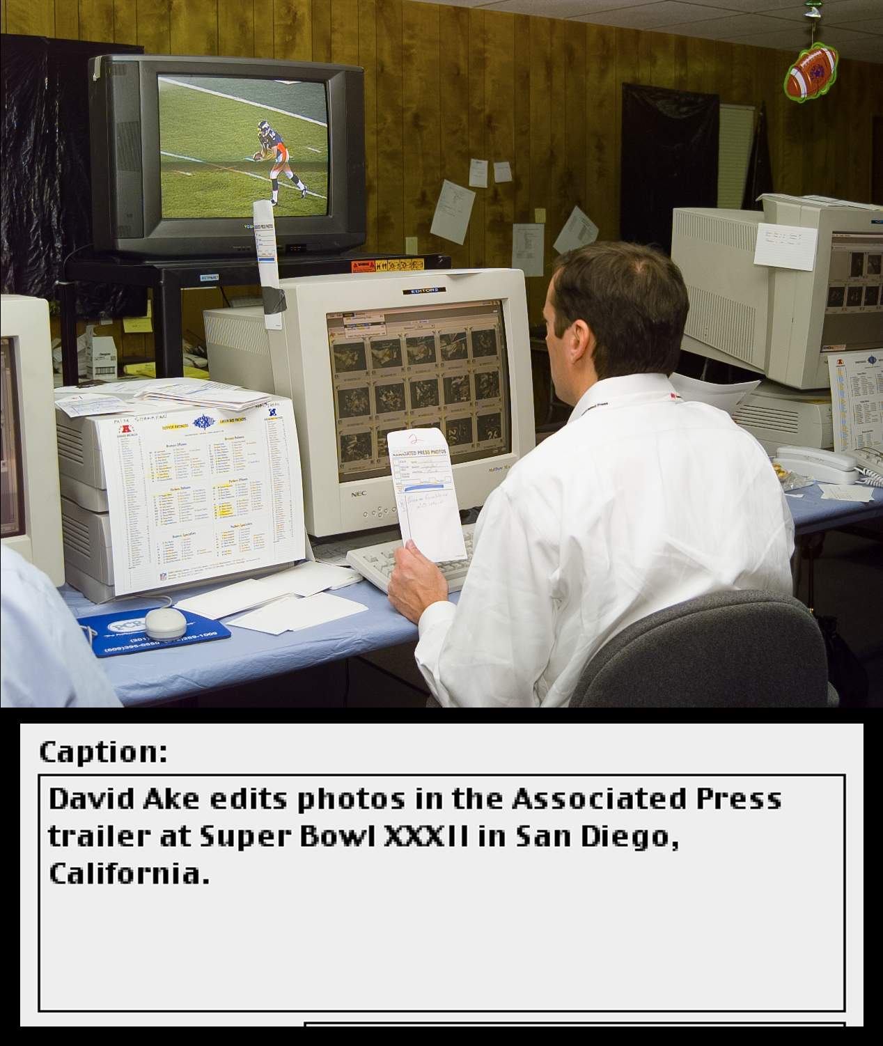 A man sits at a computer workstation and monitors an American football game on television